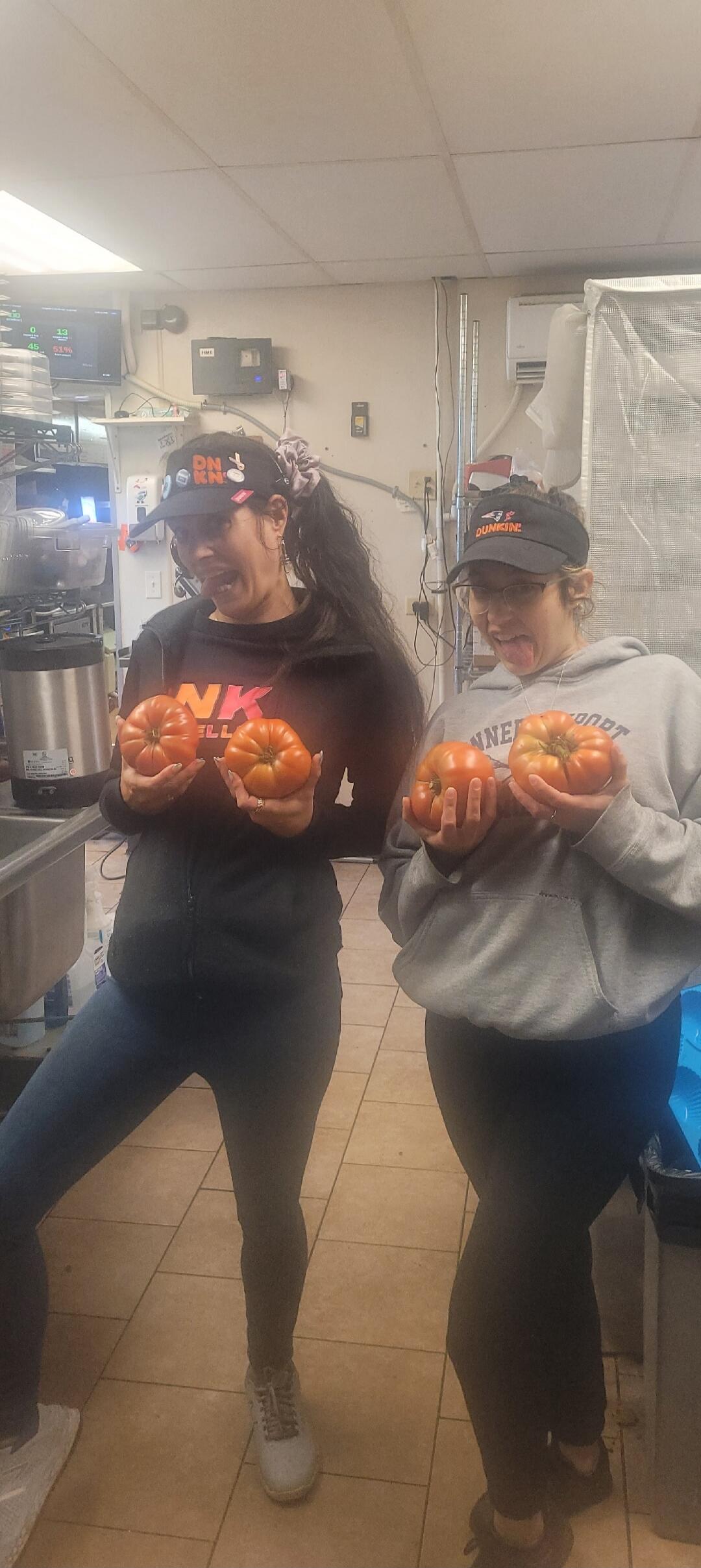 Two women in Dunkin' visors and hoodies are holding large tomatoes in front of their chests, making funny faces with their tongues sticking out. The text 'DNKN' and 'DUNKIN'' is visible on their visors, and 'NNER PORT' is partially visible on one hoodie.
