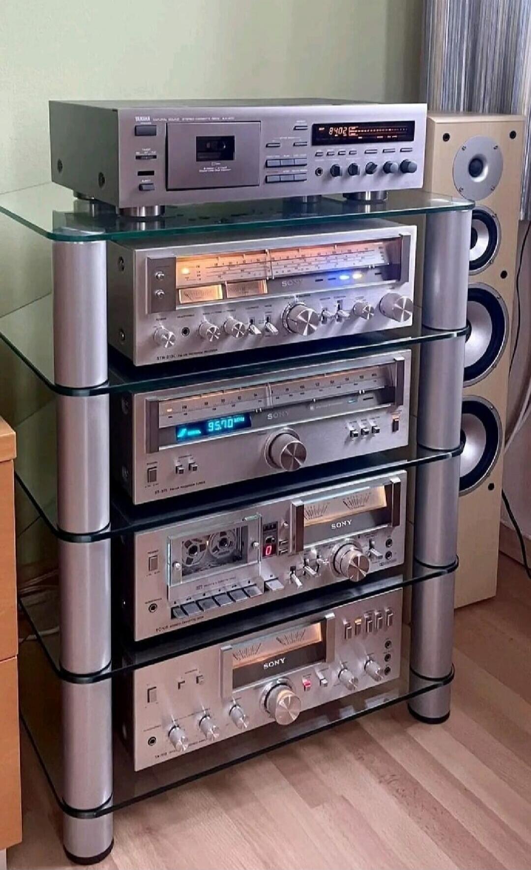 A multi-component hi‑fi stereo system with several silver amplifiers mounted in a glass-and-metal rack, accompanied by a tall floor speaker on the right.