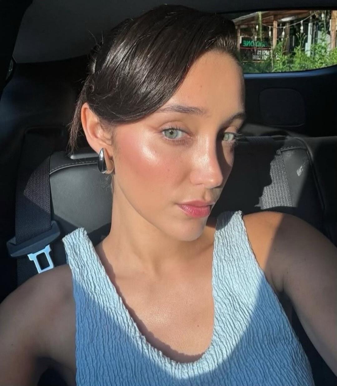 A woman in a blue sleeveless top sitting in a car, taking a selfie.