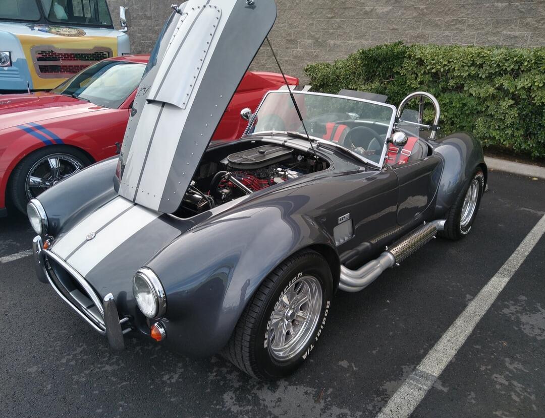 A grey vintage convertible sports car with its hood open, showing its engine. It has chrome wheels and side-exit exhaust pipes, parked in a lot beside other classic cars.