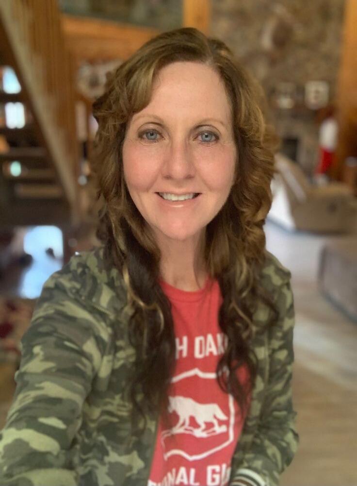A woman with long, wavy brown hair and blue eyes smiles at the camera. She is wearing a camouflage jacket over a red t-shirt that partially shows text like 'H DAK' and 'NAL G', likely referring to 'NORTH DAKOTA NATIONAL GUARD', along with a graphic of a wolf or dog. The background features a warm, indoor setting with wooden elements, including a st