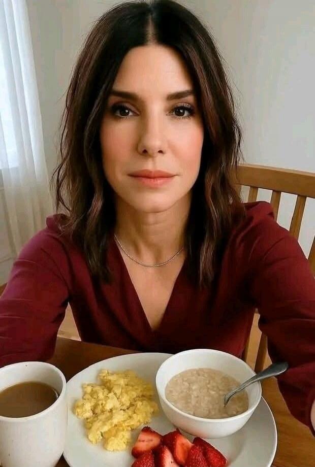 A woman with dark hair is sitting at a table, looking at the camera. In front of her is a plate with scrambled eggs, sliced strawberries, and a bowl of oatmeal. There is also a white mug with coffee.