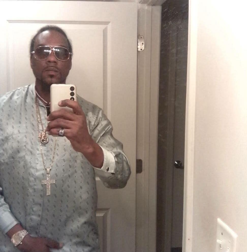 A man wearing sunglasses, a patterned shirt, and gold chains takes a mirror selfie with his phone.
