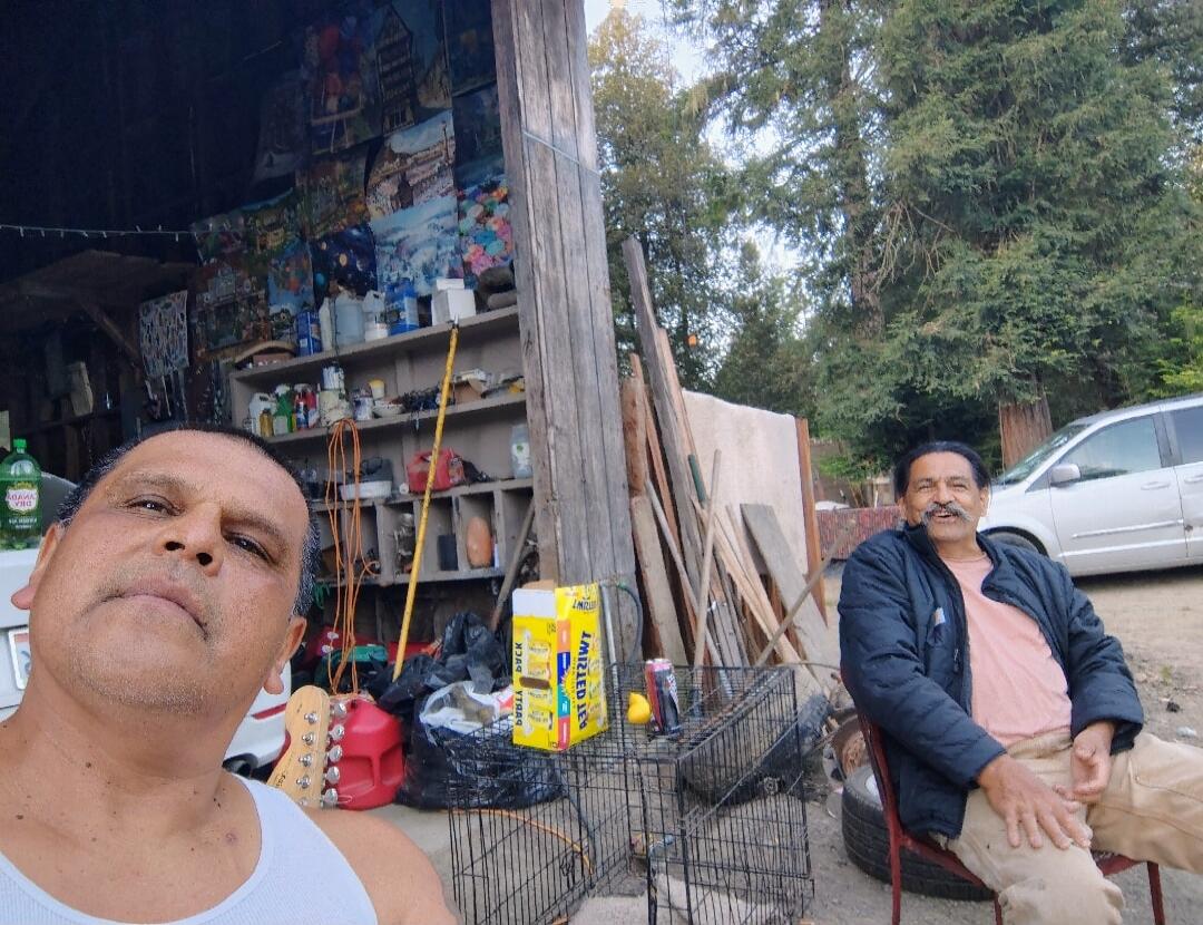 Two men are taking a selfie outdoors. One man in a white tank top is in the foreground, and another man with a mustache, wearing a dark jacket and pink shirt, is sitting in the background. Behind them is a cluttered shed with various items, including pictures on the wall, shelves, and tools. A green bottle, a red gas can, a dog crate, and a yellow 