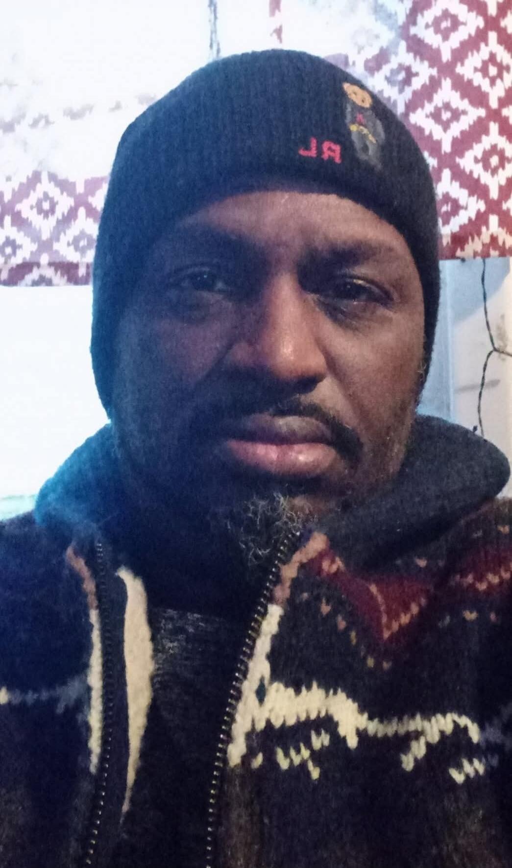 A man wearing a dark beanie with a mirrored logo and a patterned knitted jacket.