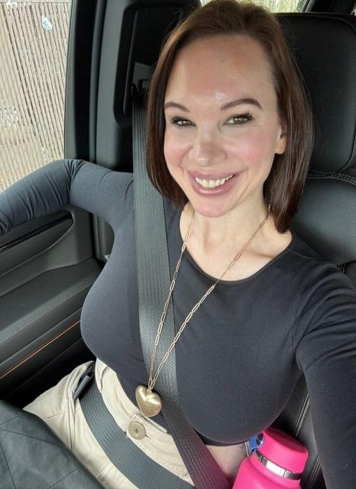 A woman is smiling and taking a selfie inside a car. She is wearing a black long-sleeved top, light-colored pants, and a seatbelt. A long necklace with a heart-shaped pendant is visible, along with a pink water bottle.