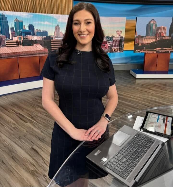 A woman, likely a news anchor, stands in a studio with a cityscape backdrop, smiling. A laptop and tablet are on a glass desk in front of her.