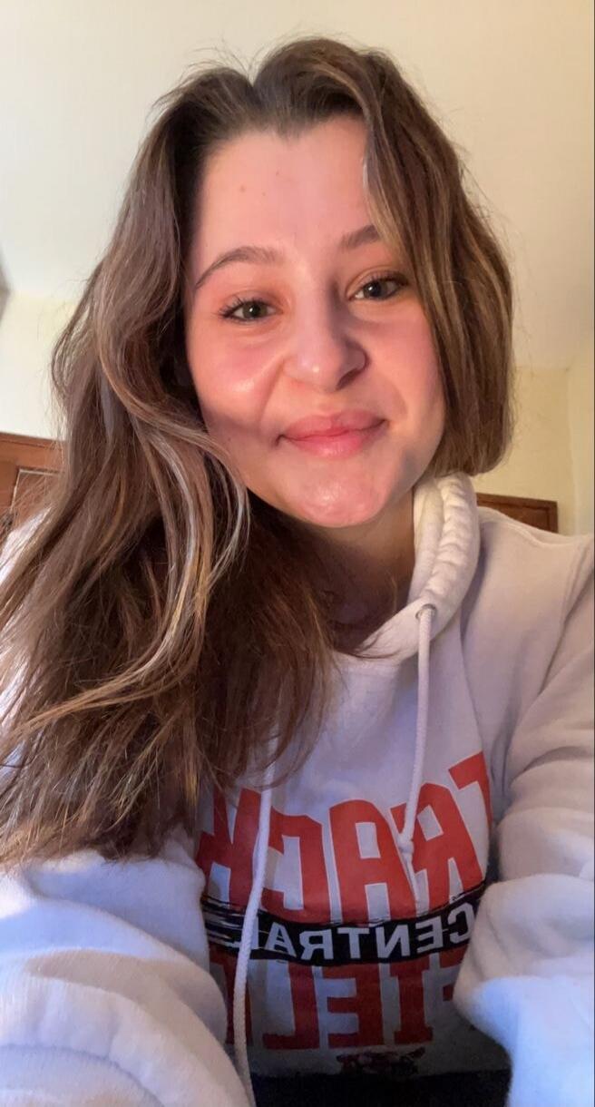 A young woman smiles at the camera, wearing a white hoodie with red text that appears to say 'TEACH'.