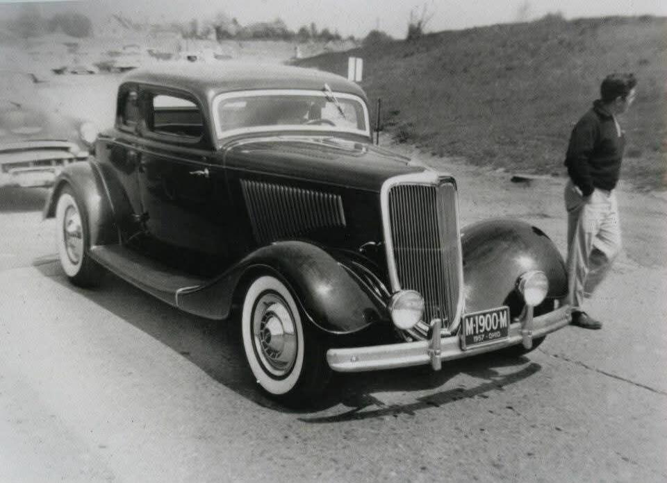 A vintage black coupe car, possibly a 1934 Ford, is parked on a paved surface. The car features whitewall tires and a prominent grille. A license plate reads 'M-1900-M 1957 - OHIO'. A man in casual attire stands next to the car on the right. The background shows a grassy hill and other vehicles in the distance.