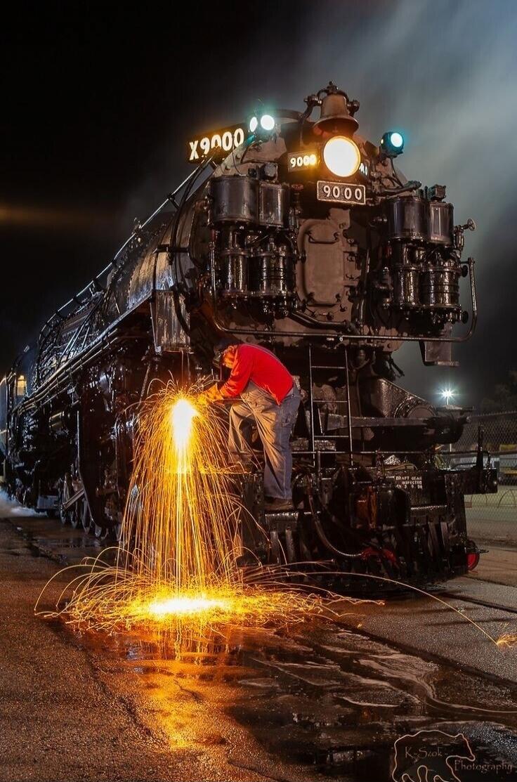 X9000 9000 – A worker in a red shirt welds on a massive black steam locomotive at night, sparks flying around. Watermark: K. S. Photography