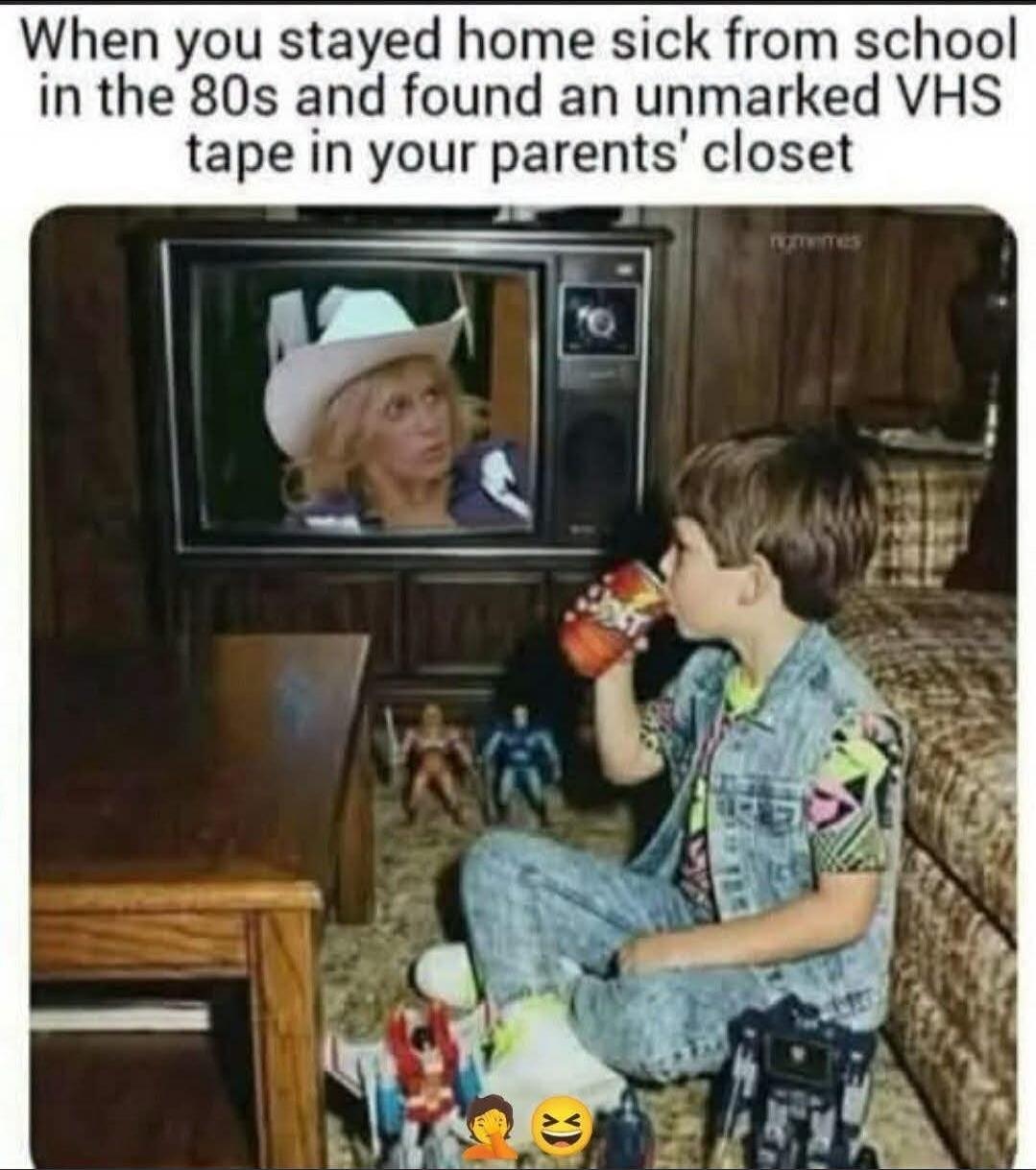 When you stayed home sick from school in the 80s and found an unmarked VHS tape in your parents' closet. A young boy is watching an old television set, where a woman in a cowboy hat is visible. The boy is drinking from a can and surrounded by toys.