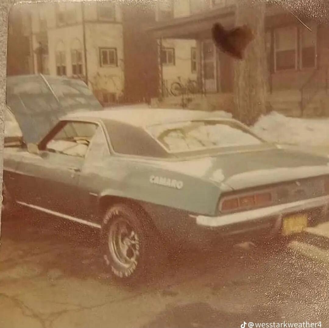 COBRA on the side of a vintage blue-green car. The photo shows a classic convertible/coupe parked on a winter street with houses in the background. There appears to be a watermark at the bottom right reading 