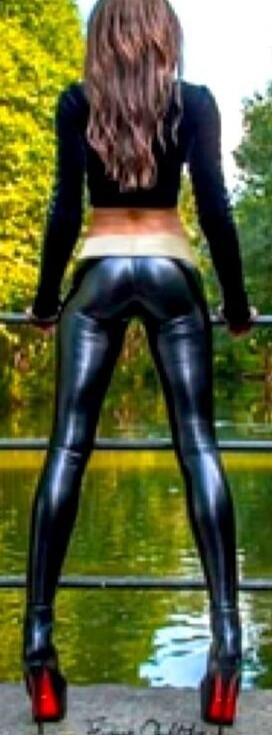 Session ID: 1059611. A person stands with their back to the camera, wearing a black long-sleeve crop top, shiny black latex leggings, and red high-heeled shoes, outdoors by a railing near water.