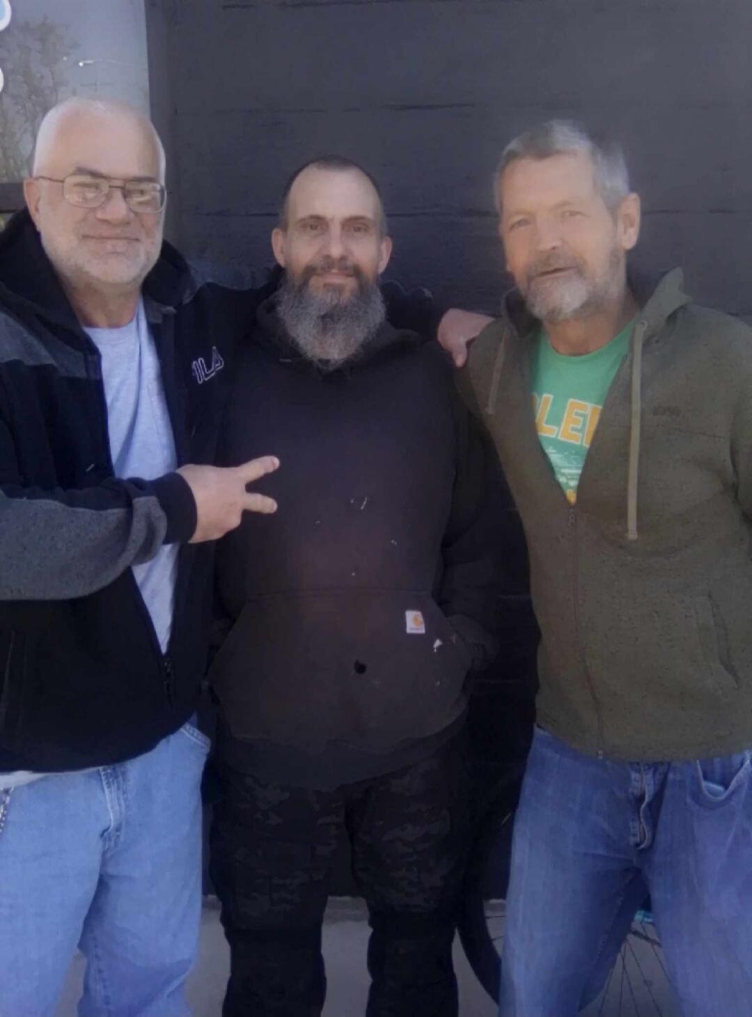 Three men are standing together, smiling. One man on the left is wearing glasses and a black and grey jacket with 'ILA' visible. The man in the middle has a beard and is wearing a black hoodie with a small Carhartt logo on the pocket. The man on the right has a beard and is wearing a green t-shirt with 'LE' visible under an olive green zip-up hoodi