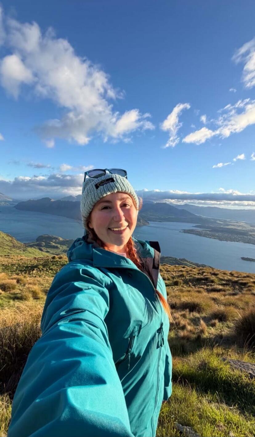 A young woman is taking a selfie on a mountain top with a lake and mountains in the background.