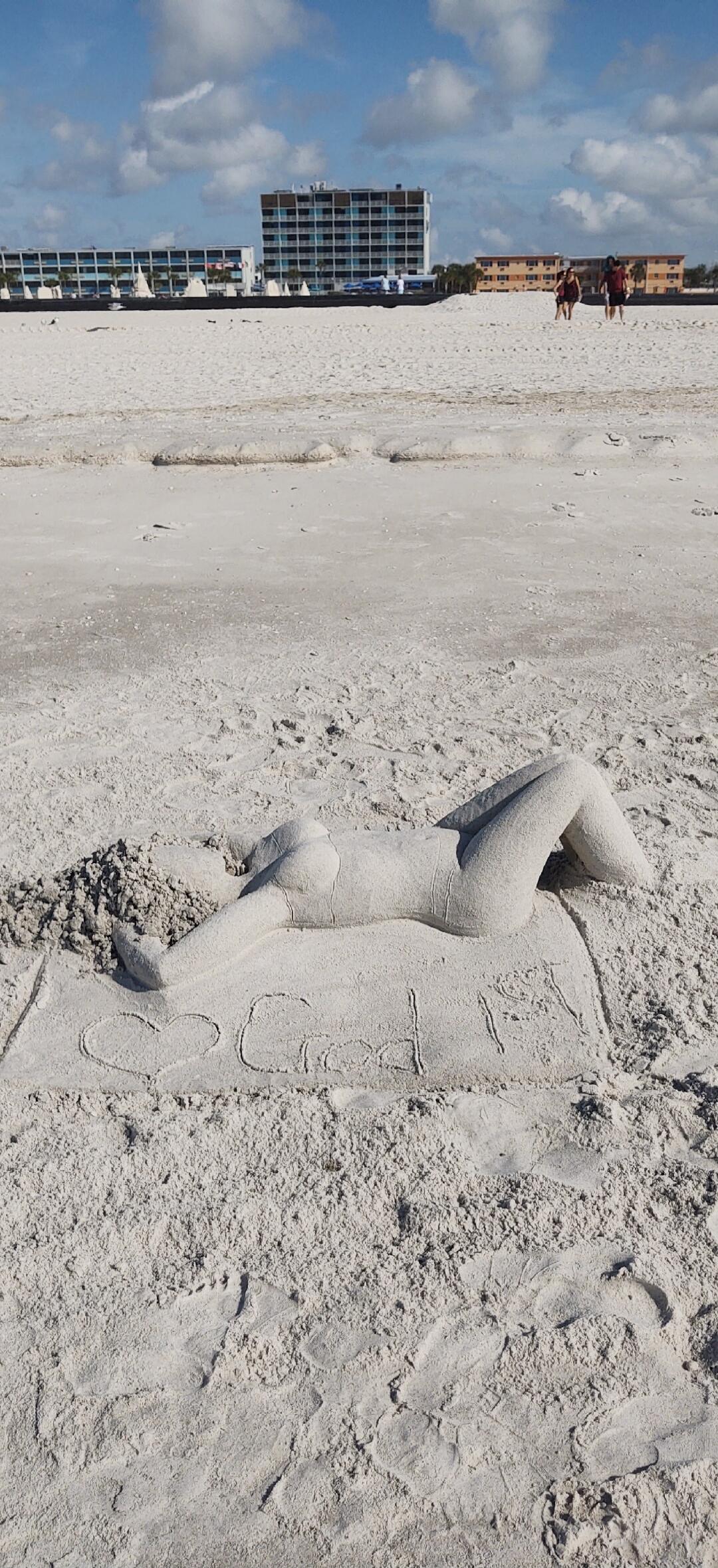 A sand sculpture of a reclining female figure on a beach. The word 'Good' and a heart shape are drawn in the sand next to it.