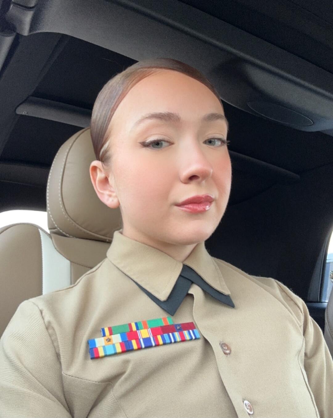 Portrait of a young person in a tan military uniform with ribbons on the chest, seated in a vehicle.