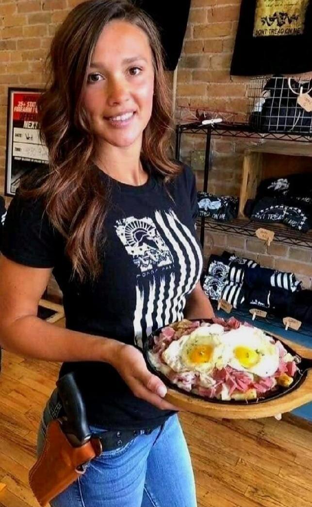 A woman wearing a black t-shirt with a design (possibly flag) holds a plate with fried eggs and slices of meat in a shop or cafe setting.