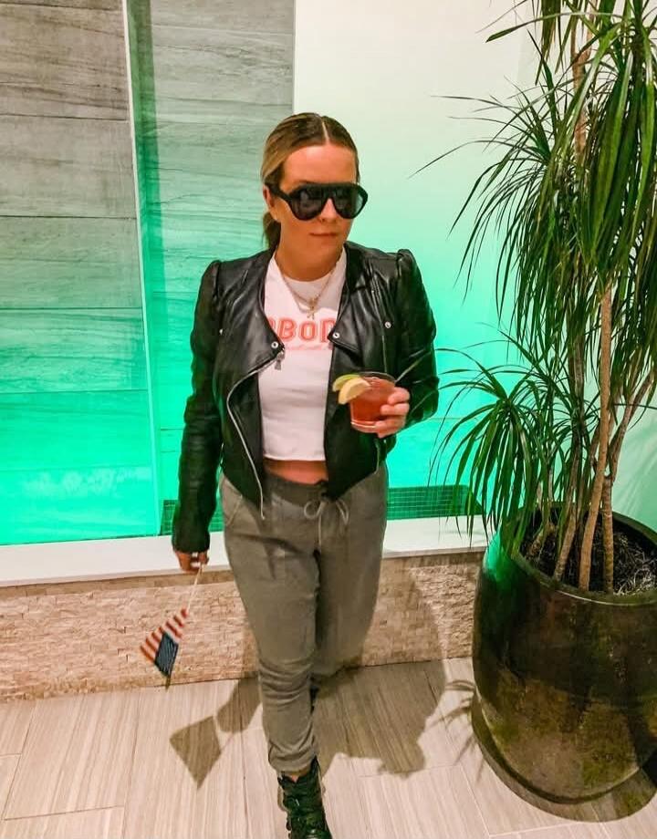 A person wearing a black leather jacket over a white cropped top with red lettering that appears to say 'BOY'. They are also wearing gray joggers, black boots, and large sunglasses. They are holding a cocktail with a lime wedge in one hand and a small American flag in the other. Green lighting in the background.
