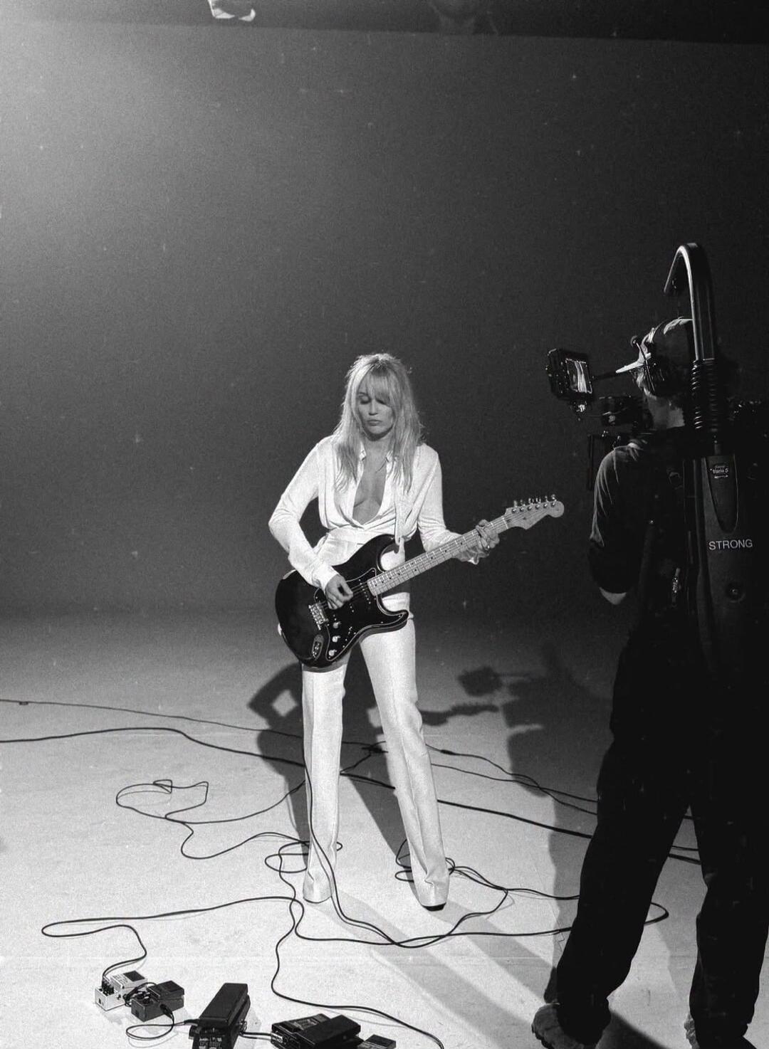 A woman with long blonde hair is playing an electric guitar in a studio setting. She is wearing a white shirt with a deep V-neck and white pants. A cameraman is filming her. The word 'STRONG' is visible on the cameraman's equipment.