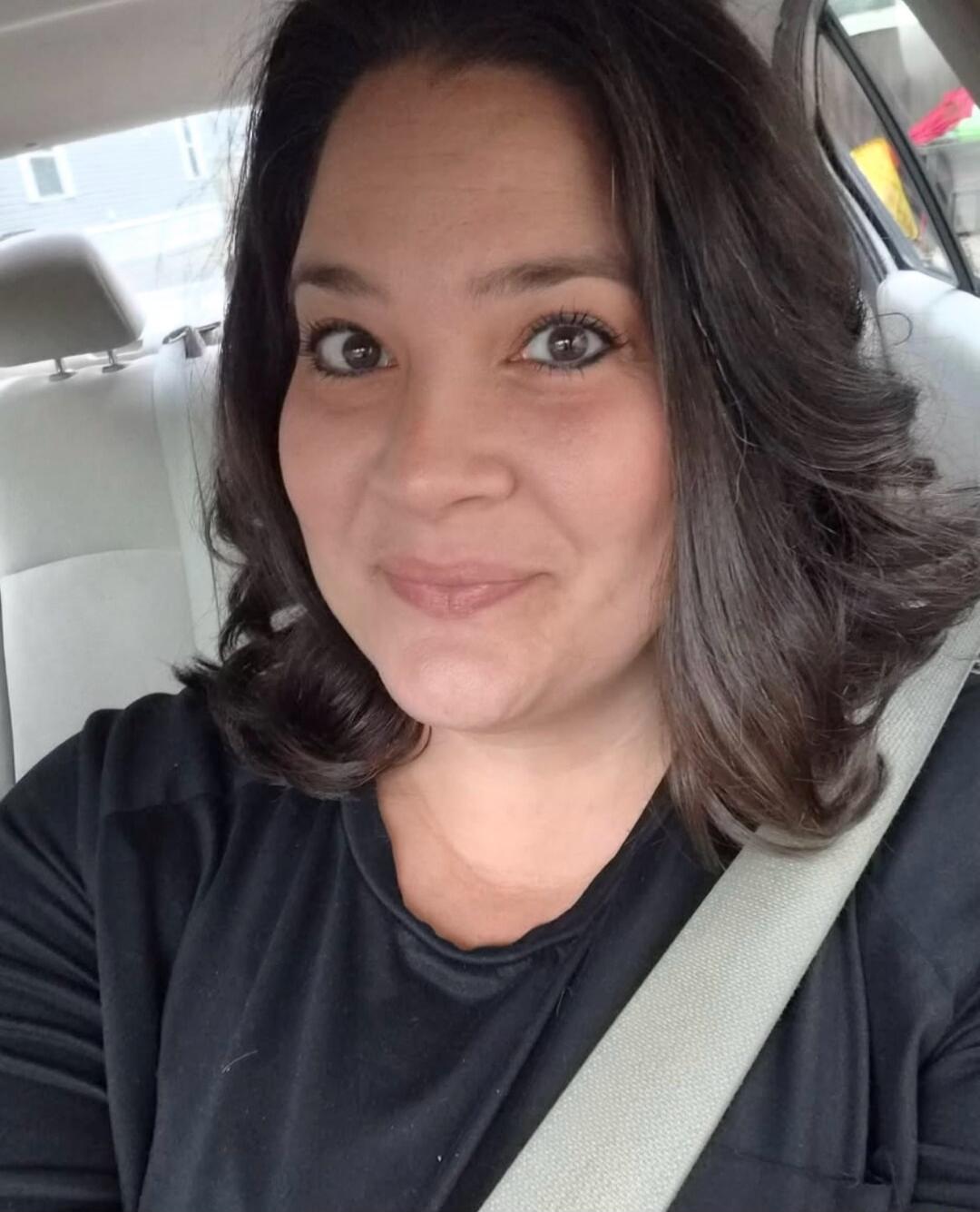 A woman sitting in a car, wearing a black top, smiling at the camera.