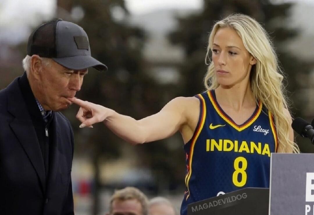 A woman in an Indiana basketball jersey with number 8 is touching an older man's mouth with her finger. The man is wearing a cap and a dark jacket. The woman has a serious expression. Text on the podium: MAGADEVILDOG.