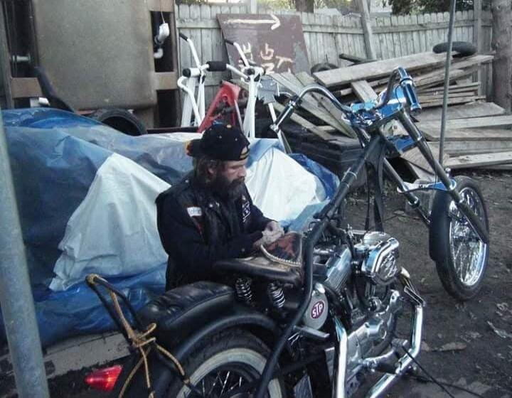 A bearded man in a cap and vest is sitting next to a custom motorcycle (chopper), possibly working on it or examining something in his hands. The motorcycle has an 'STP' logo on its engine. The background shows a cluttered outdoor area with tarps and wood.