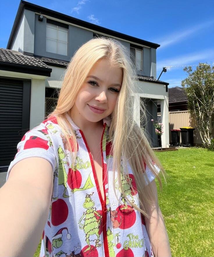 A young woman takes a selfie outdoors in front of a modern house. She is wearing a light-colored shirt with a Grinch pattern and has long blonde hair.