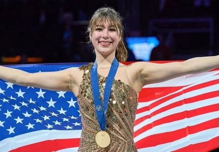 A female athlete on stage with a gold medal, standing in front of an American flag with arms outstretched in celebration.