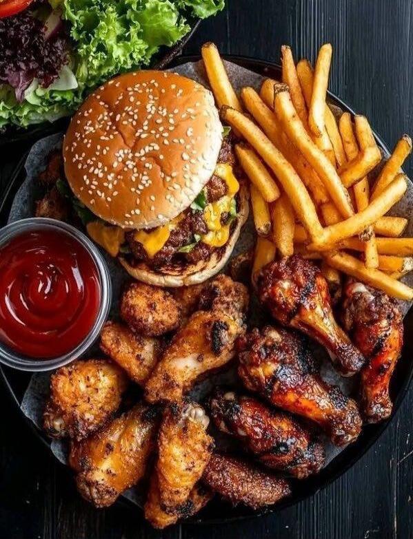 A plate of food featuring a sesame bun burger with cheese, crispy chicken wings, crispy french fries, and bite-sized fried chicken nuggets, with a side of ketchup and leafy greens in the background.