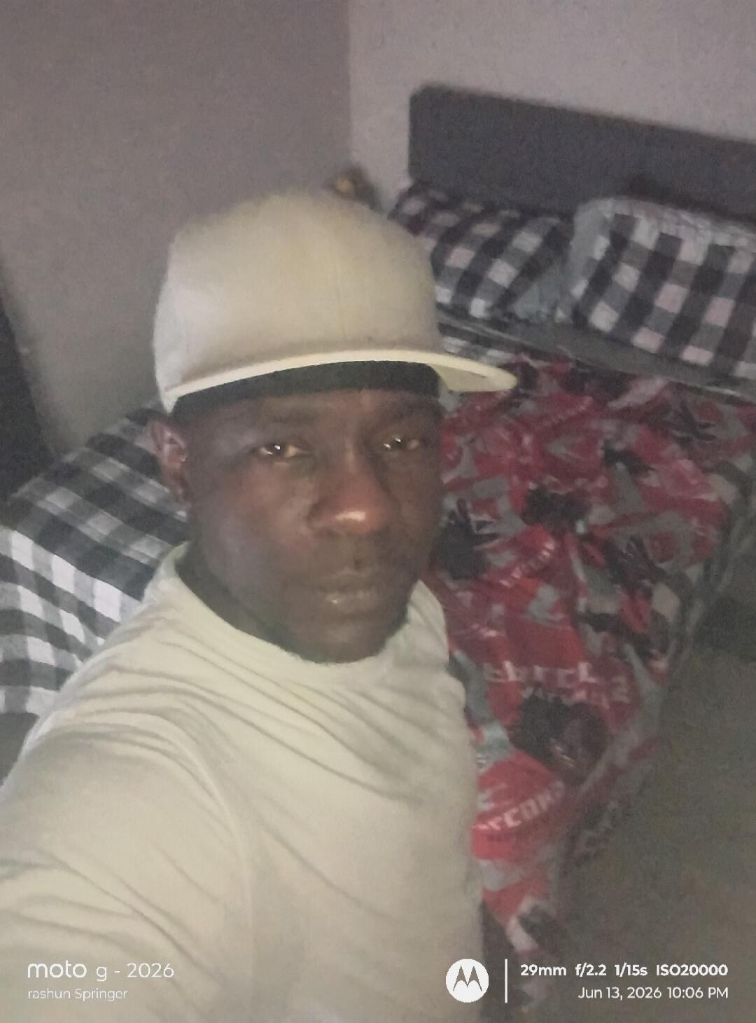 moto g - 2026 rashun Springer 29mm f/2.2 1/15s ISO20000 Jun 13, 2026 10:06 PM. A man in a light t-shirt and a baseball cap taking a selfie in a room with a bed in the background.