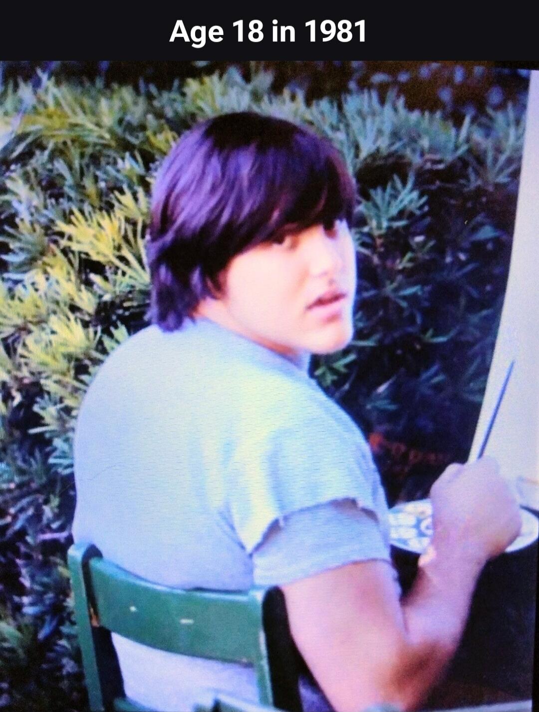 Age 18 in 1981. A young person with dark hair is sitting on a green chair, looking back over their shoulder. They appear to be painting, holding a brush and a palette.
