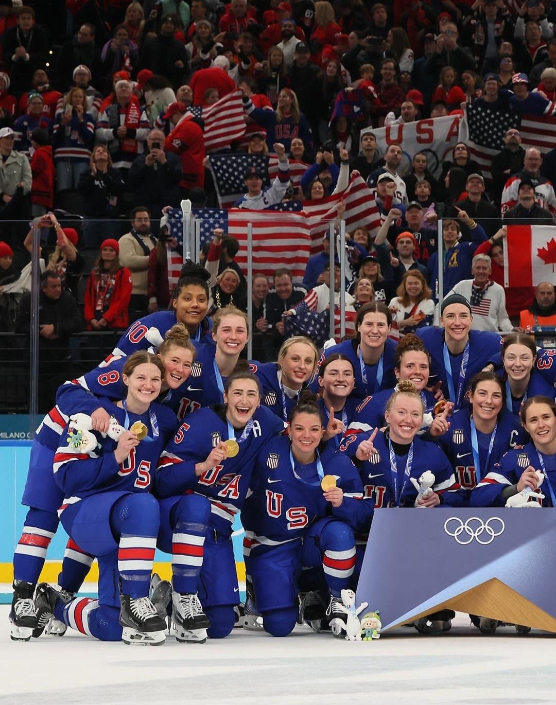 US women's ice hockey team posing with medals on the ice, surrounded by cheering fans holding American flags at the Olympics.