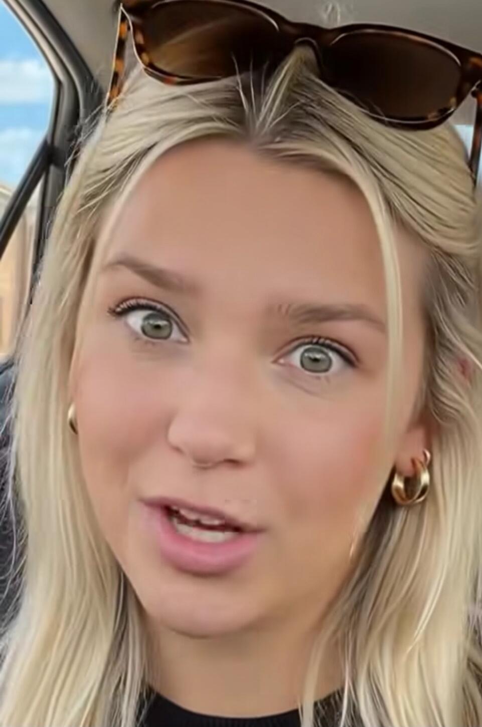 A young blonde woman with wide eyes and an open mouth, possibly speaking or reacting with surprise, in a car.