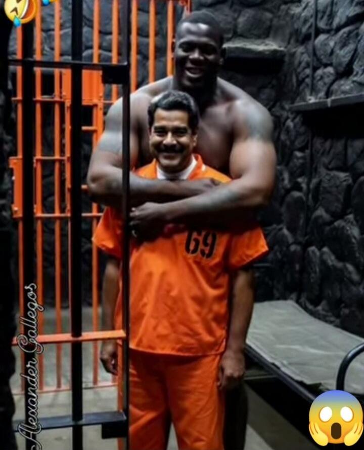 Two men; one in an orange prison jumpsuit with the number 69, being hugged from behind by a much taller man who is shirtless, standing beside a barred orange gate.