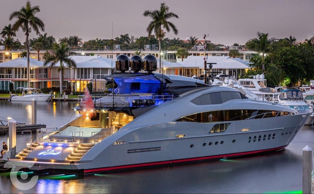 A luxurious modern yacht, possibly named 'Bellami', docked in a marina at dusk, with upscale houses and palm trees in the background. The yacht features blue accents and illuminated steps.