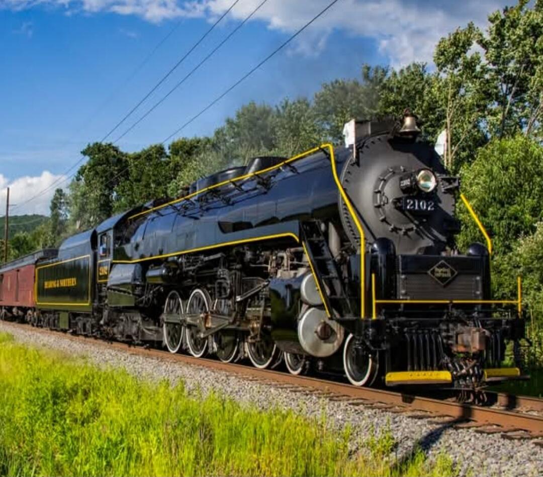 A classic black steam locomotive with yellow trim on a railway track, pulling a tender car through a green, rural landscape.