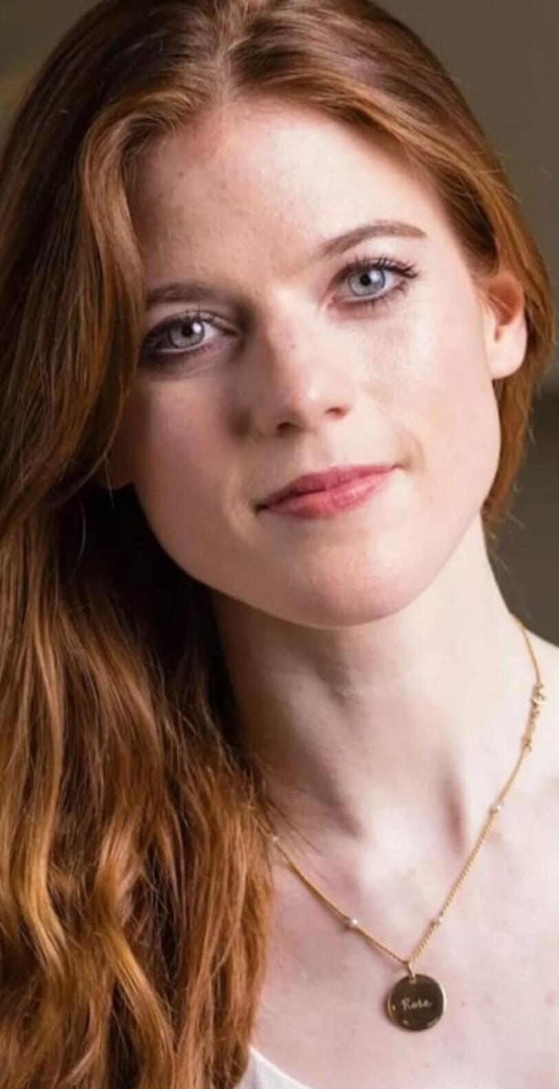 A woman with long reddish-brown hair and blue eyes, wearing a necklace with a 'Rose' pendant.