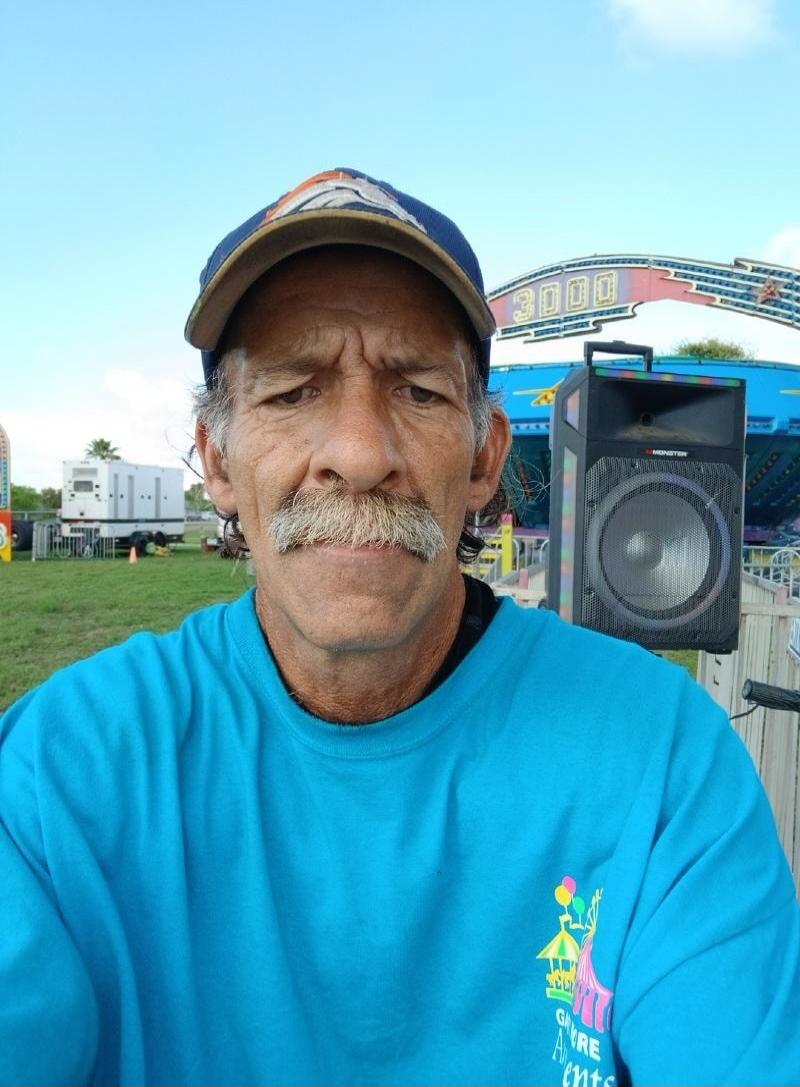 A man with a mustache and a cap, wearing a blue t-shirt with 'GORE Events' logo, stands outdoors. In the background, a carnival ride with '3000' written on it and a 'MONSTER' brand speaker are visible.