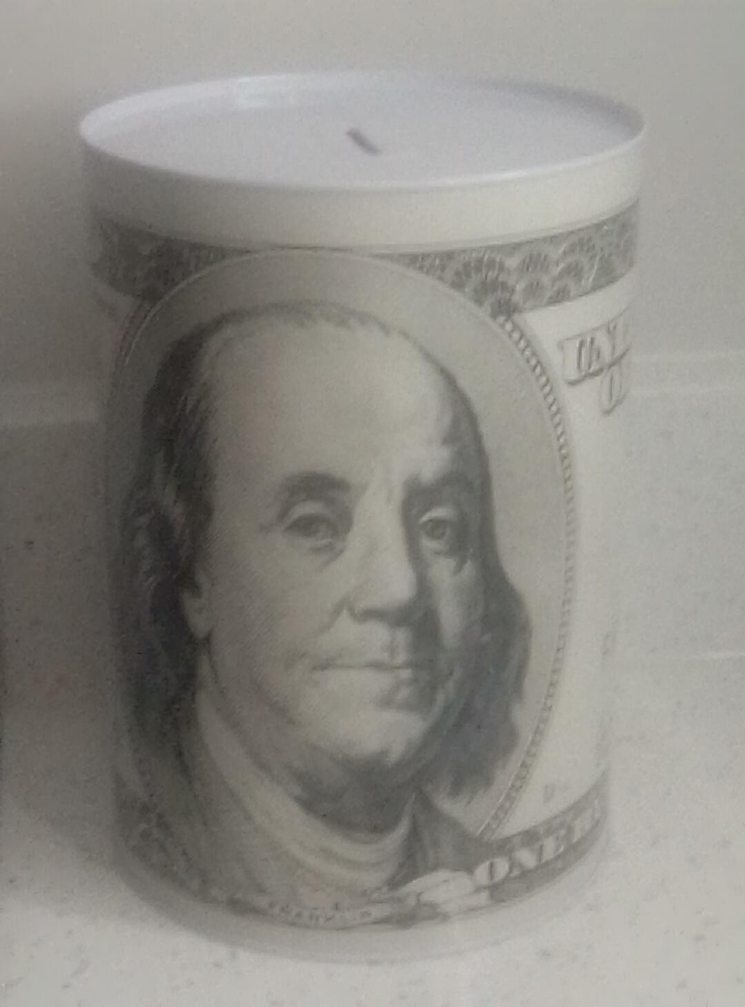 A cylindrical money box designed to resemble a US 100 dollar bill, featuring the portrait of Benjamin Franklin and text like 'ONE HUNDRED DOLLARS' and 'THE UNITED STATES OF AMERICA'. There is a slot on top for inserting coins or bills.