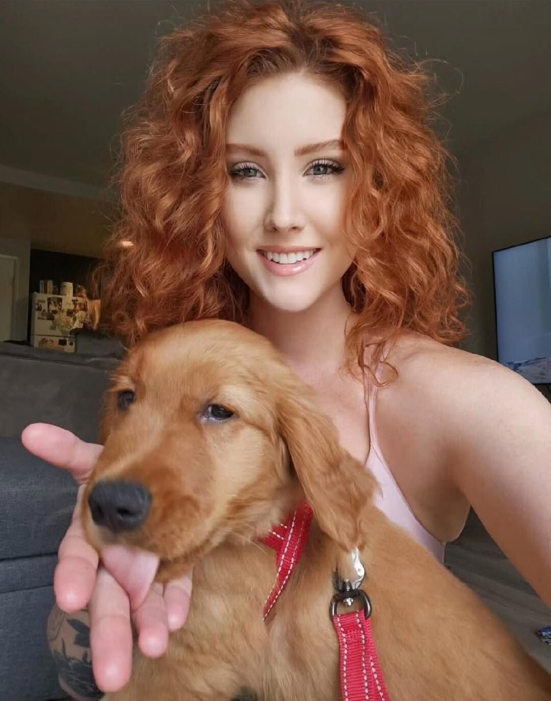A woman with curly red hair smiling and holding a young golden retriever wearing a red collar.