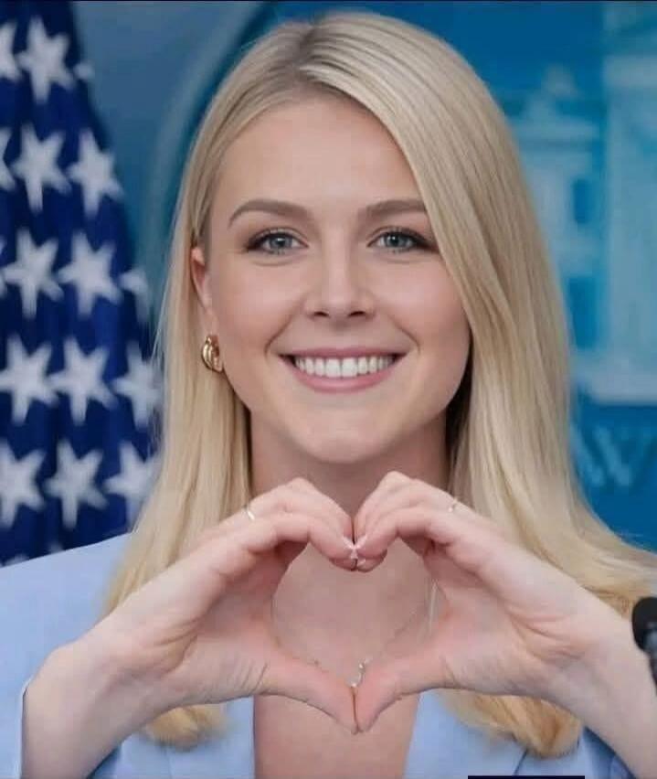 A woman is making a heart shape with her hands in front of her chest. She is smiling and looking at the camera. There is an American flag in the background.
