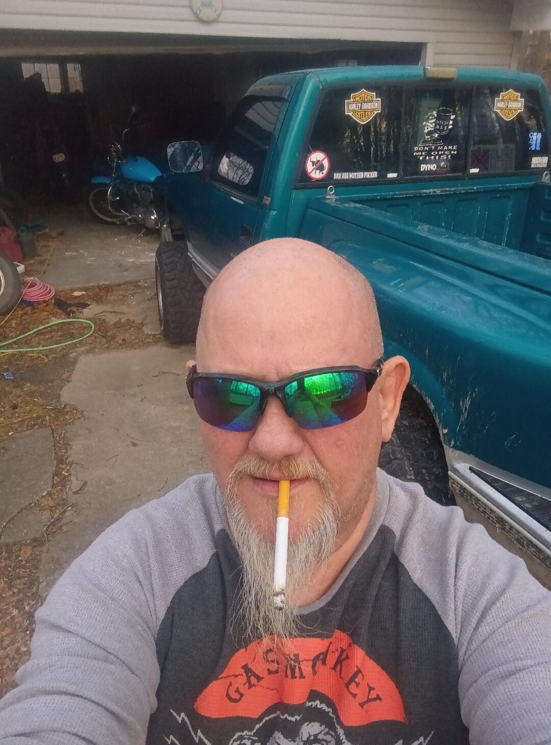 A man with a beard and sunglasses holds an unlit cigarette in his mouth. He is wearing a 'GASMONKEY' shirt. In the background, there is a teal pickup truck with 'HARLEY-DAVIDSON CYCLES' stickers, a 'BAD ASS MOTHER FUCKER' sticker, and a 'DON'T MAKE ME OPEN THIS!' sticker with a skull graphic, as well as a 'DYNO' sticker. A blue motorcycle is visibl