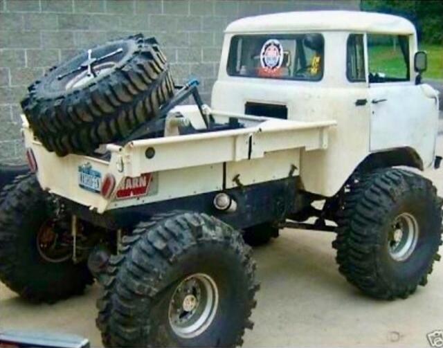 A vintage white pickup-style vehicle with massive off-road tires, a spare tire mounted in the bed, and a raised suspension. There is a small sticker on the back of the cab and a large tire strapped in the bed.