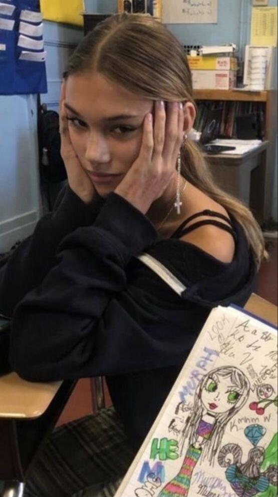 A young woman sits in a classroom with her hands pressed to the sides of her face, wearing a black off-shoulder top and earrings. She has a colorful drawing hanging in front of her, showing a illustrated girl with green eyes and long hair.