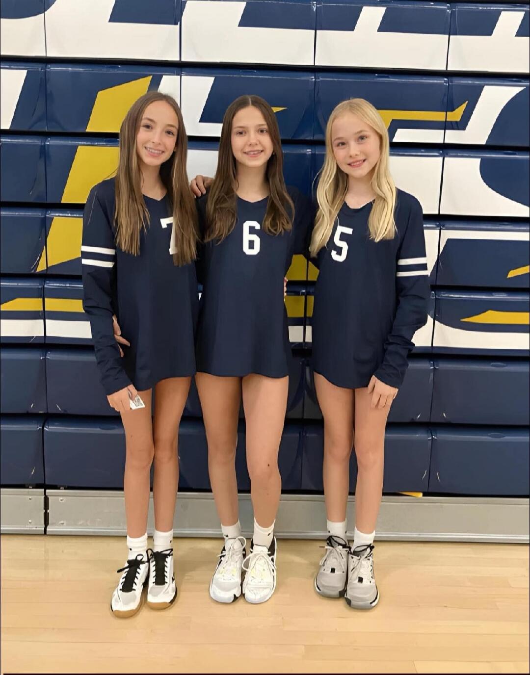 Three young girls wearing dark blue long-sleeve sports shirts with numbers 7, 6, and 5, standing in front of a blue and yellow wall.