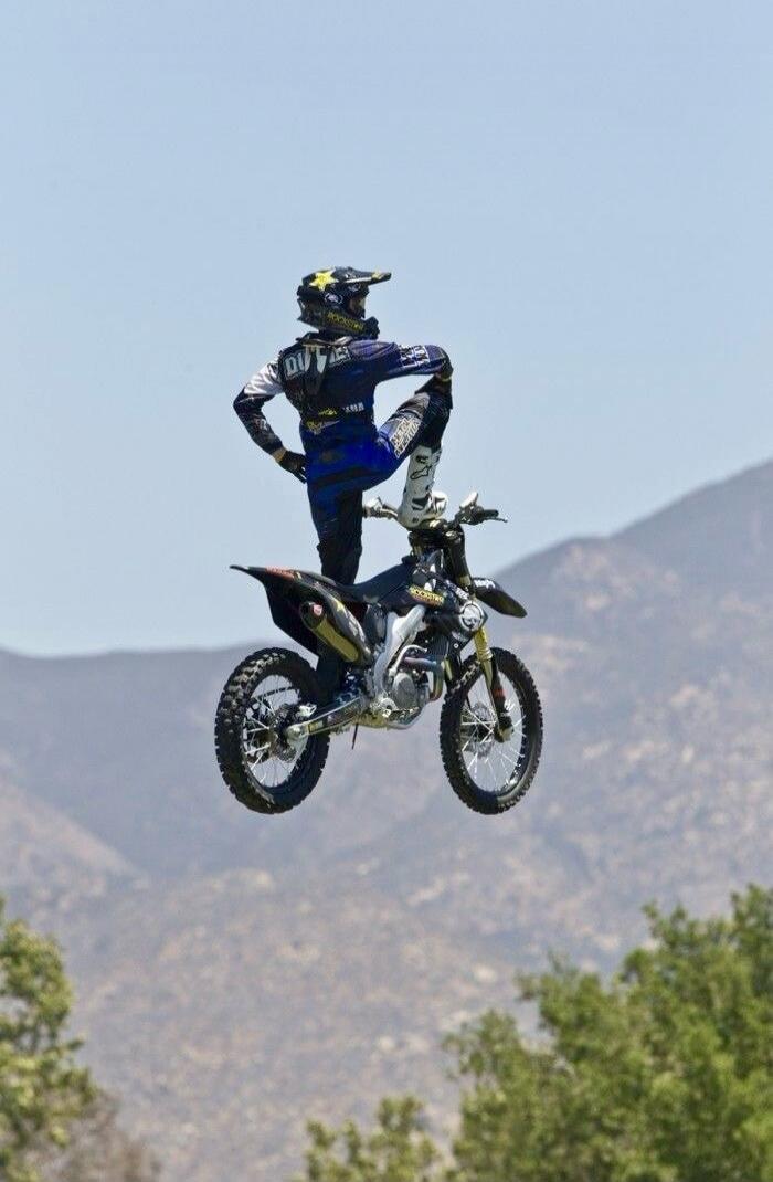 A motocross rider wearing blue gear is mid-air on a dirt bike performing a jump, with mountains in the background.