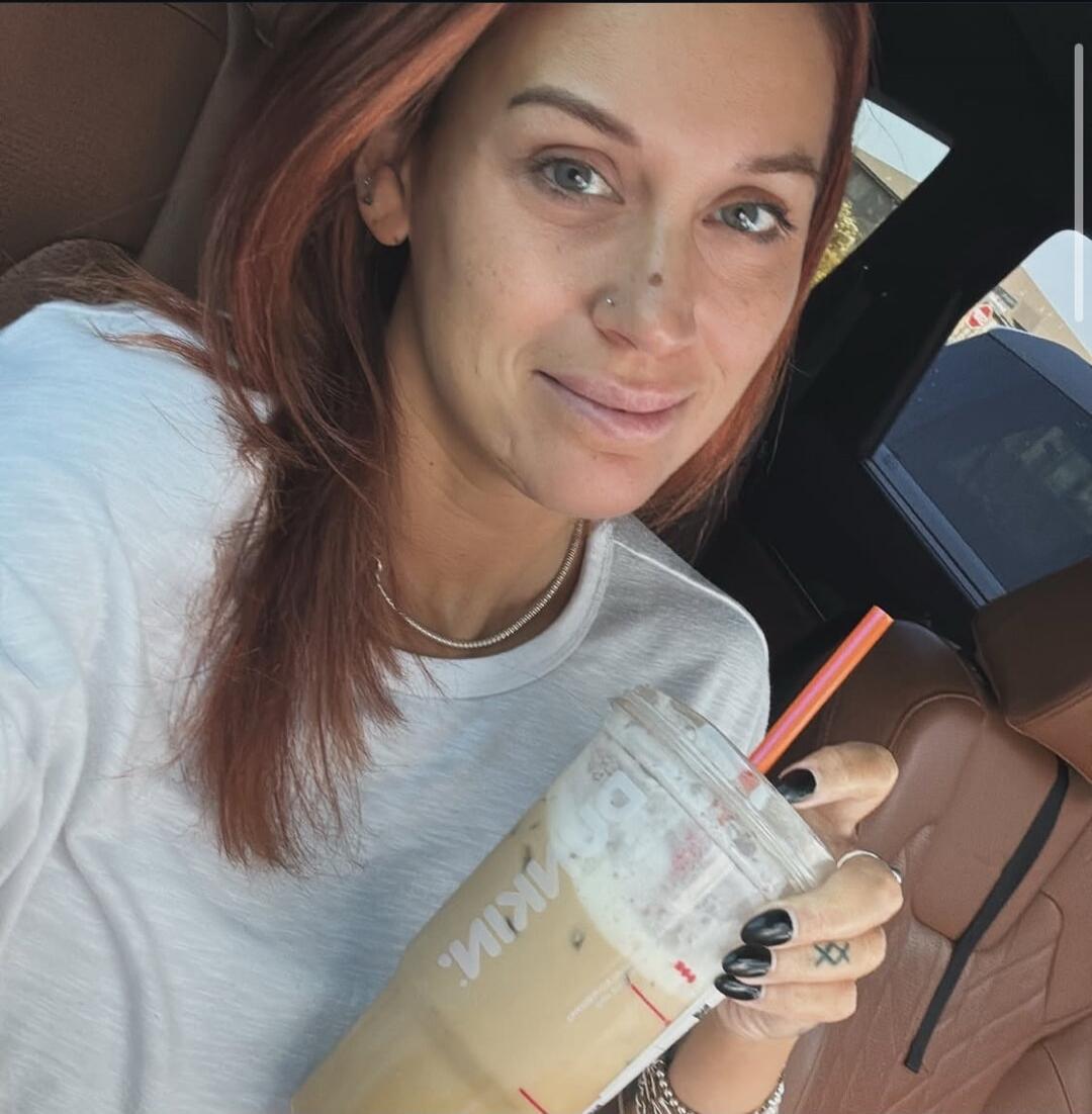 A woman with reddish-brown hair in a white t-shirt is in a car, holding a large iced coffee cup from Dunkin'. She is smiling at the camera.