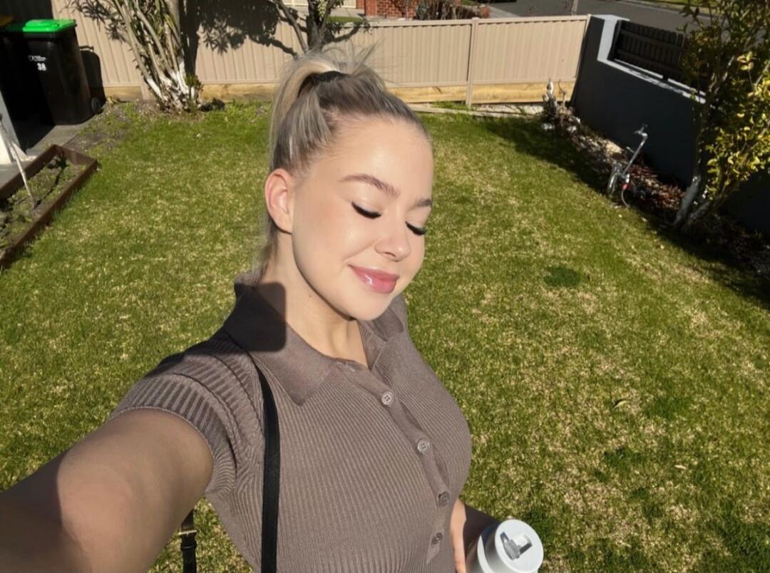 A young woman with blonde hair tied in a ponytail is taking a selfie outdoors in a sunny garden. She is smiling with her eyes closed, enjoying the sun, and holding a white cup. She is wearing a brown ribbed polo shirt.