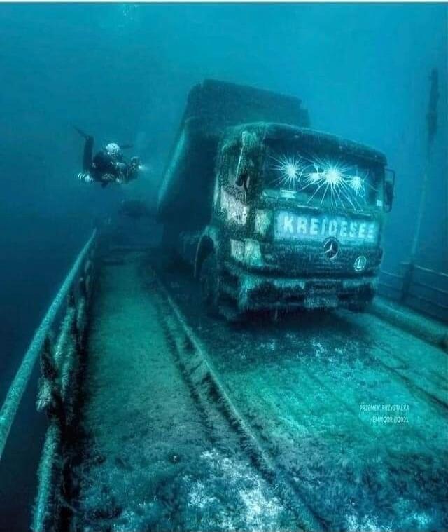 KREIDESEE PRZEMEK PRZYSTALKA HEMMOOR ©2021. An underwater scene featuring a diver exploring a large, submerged truck at the bottom of a clear blue lake.